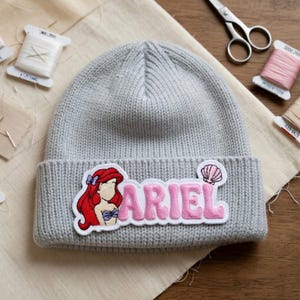 May include: A gray knit beanie featuring a patch with the name "ARIEL" in pink letters and an illustration of Ariel from The Little Mermaid. The patch includes a seashell graphic. The beanie is on a beige fabric.