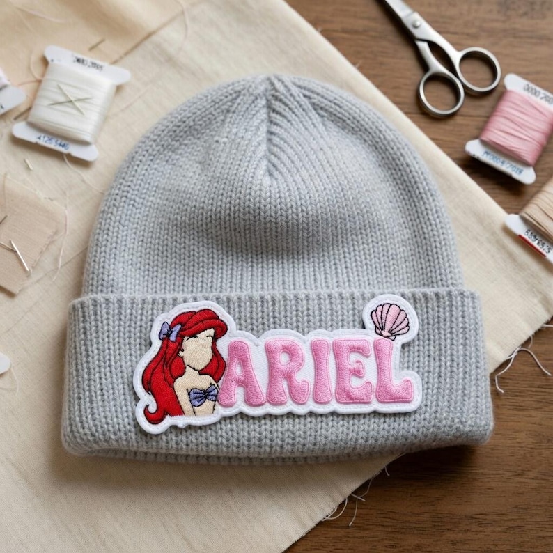May include: A gray knit beanie featuring a patch with the name "ARIEL" in pink letters and an illustration of Ariel from The Little Mermaid. The patch includes a seashell graphic. The beanie is on a beige fabric.