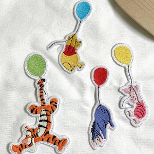 Könnte beinhalten: Gestickte Aufnäher mit Winnie-Puuh-Figuren: Winnie Puuh, Tigger, Eeyore und Ferkel, jeweils mit einem bunten Ballon. Die Aufnäher sind weiß umrandet und haben leuchtende Farben.