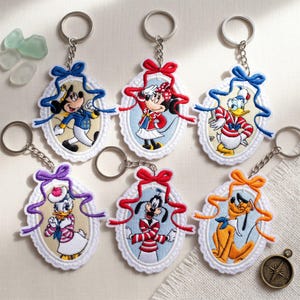 May include: Six oval embroidered keychains featuring Disney characters Mickey Mouse, Minnie Mouse, Donald Duck, Daisy Duck, Goofy, and Pluto. Each character is framed in a white scalloped border with a colorful ribbon bow at the top and a metal ring.