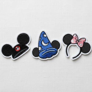 Puede incluir: Tres parches bordados con icónicas orejas de ratón. Un parche representa un sombrero negro con un emblema rojo y blanco, otro un sombrero de mago azul con estrellas y una luna creciente, y el último una diadema con un lazo rosa.