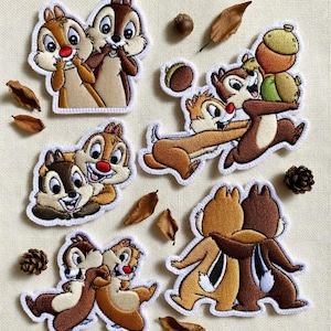 Op de afbeelding: Geborduurde patches met Chip en Dale, de Disney-chipmunk duo, in verschillende poses. De patches zijn bruin, beige en wit met rode neuzen, omringd door bladeren, eikels en dennenappels op een crèmekleurige achtergrond.