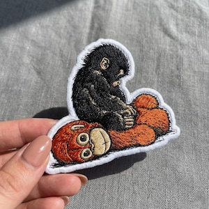 May include: Embroidered patch featuring a black monkey sitting on top of a red-orange monkey. The patch has a white border. The monkey's fur is detailed with various shades of black and brown. The red-orange monkey has a smiling face.