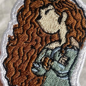 May include: Embroidered patch featuring a cartoon illustration of a woman with long, wavy auburn hair. She wears a green dress and has a black and green headpiece. The patch has a white border.