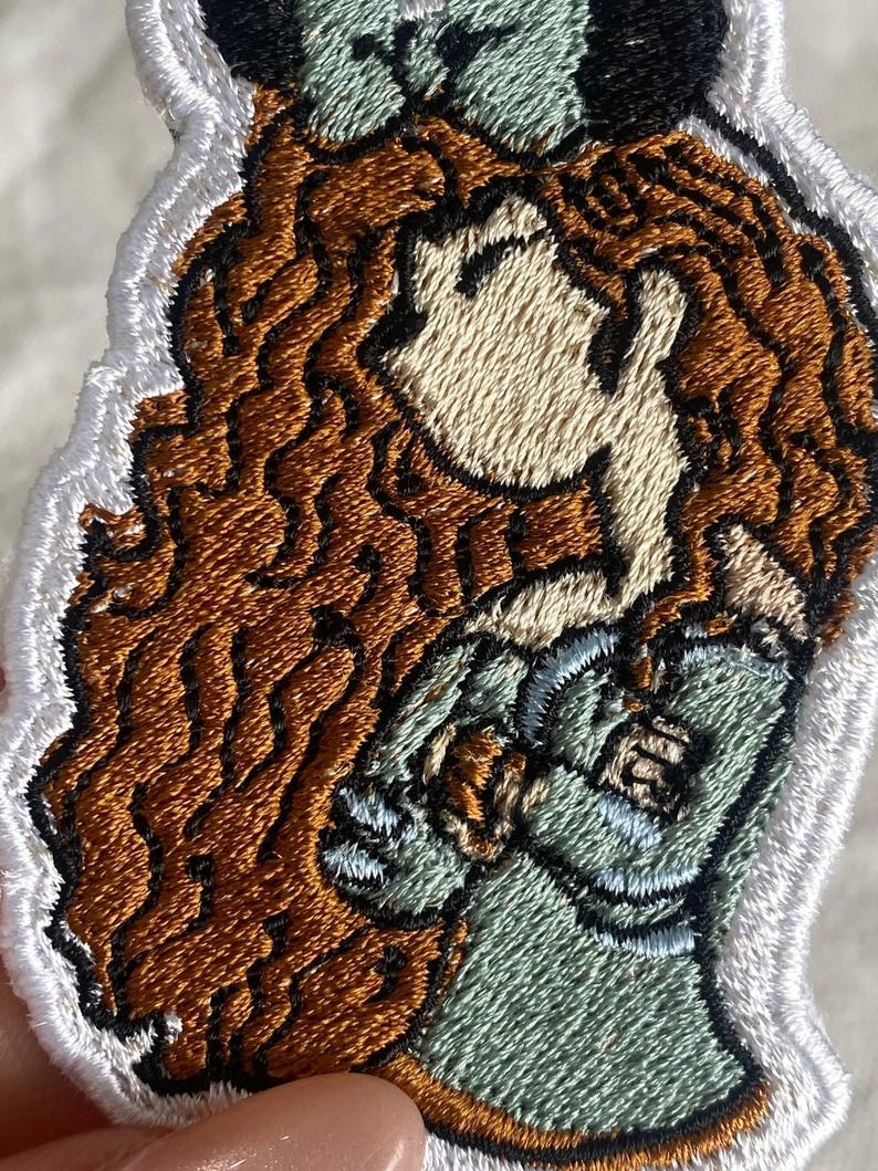May include: Embroidered patch featuring a cartoon illustration of a woman with long, wavy auburn hair. She wears a green dress and has a black and green headpiece. The patch has a white border.