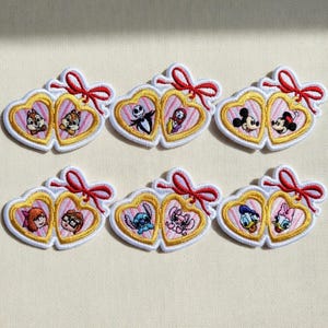 Op de afbeelding: Geborduurde patches met paren stripfiguren in hartvormige frames. Elk hart is goudkleurig omlijnd met een witte rand en een rode strik. Personages zijn onder meer Knabbel en Babbel, Jack Skellington en Sally, Mickey en Minnie Mouse.