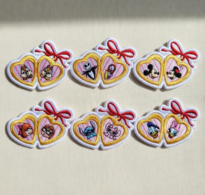 Op de afbeelding: Geborduurde patches met paren stripfiguren in hartvormige frames. Elk hart is goudkleurig omlijnd met een witte rand en een rode strik. Personages zijn onder meer Knabbel en Babbel, Jack Skellington en Sally, Mickey en Minnie Mouse.