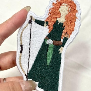 Op de afbeelding: Geborduurde patch met een personage met rood haar, gekleed in een groene jurk en met een boog en pijl. De patch heeft een witte rand en is geschikt voor kleding of accessoires.