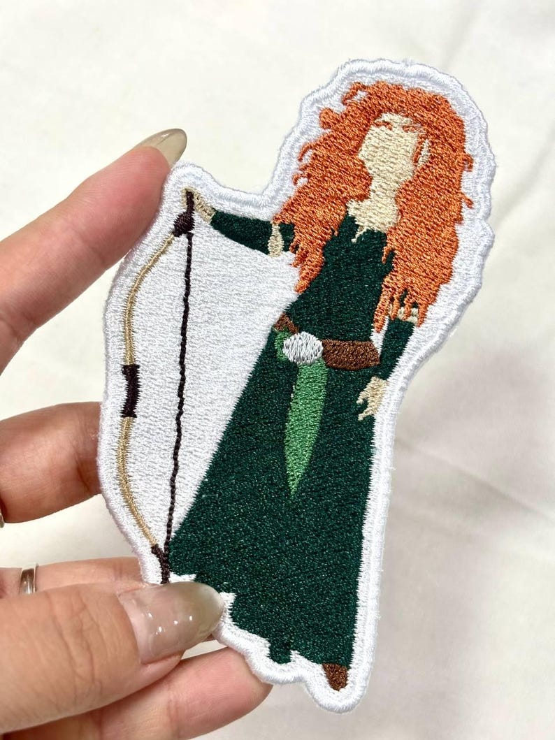 Op de afbeelding: Geborduurde patch met een personage met rood haar, gekleed in een groene jurk en met een boog en pijl. De patch heeft een witte rand en is geschikt voor kleding of accessoires.
