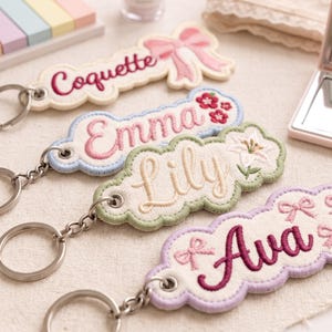 May include: Four embroidered fabric keychains with scalloped edges and metal rings. Each features a name in cursive script: Coquette with a pink bow, Emma with red flowers, Lily with a white lily flower, and Ava with small pink bows.