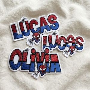 Könnte beinhalten: Drei gestickte Aufnäher mit den Namen LUCAS und OLIVIA in einer rot-blauen Schriftart mit Spinnennetz-Muster. Jeder Aufnäher enthält eine kleine Comic-Spinnenfigur und eine Netzgrafik, umrandet von einer weißen Kontur.