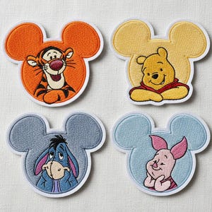 May include: Four embroidered patches featuring Disney characters. Tigger, in orange and black, Winnie the Pooh in yellow and red, Eeyore in blue, and Piglet in pink, are set within a Mickey Mouse head shape with a white border.