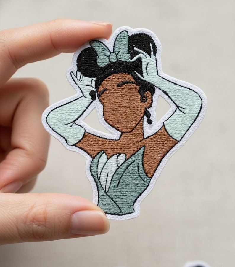 May include: Embroidered patch featuring a cartoon illustration of a woman with dark skin, wearing a light green dress, gloves, and a bow. The patch has a white border and is held up by a hand.