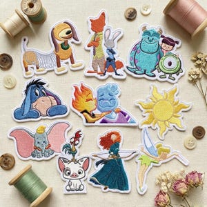 Könnte beinhalten: Eine Sammlung gestickter Aufnäher mit verschiedenen Zeichentrickfiguren, darunter Eeyore, Dumbo und Sully. Die Aufnäher sind farbenfroh und detailliert gestickt. Sie liegen auf hellem Untergrund, mit Nähutensilien und Knöpfen.