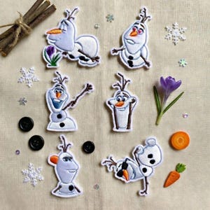 May include: Embroidered patches featuring the snowman Olaf from the movie Frozen. The patches are white, blue, and orange, with black outlines. The image also includes buttons, a flower, and a carrot slice.