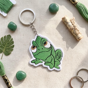 以下が含まれることがあります： 大きな白い目と黒い輪郭が刺繍された、緑色のカメレオンのキーホルダー。カメレオンは短いチェーンで銀色の金属製二重リングに取り付けられています。デザインには白い縁取りがあり、表面は質感のある布地でできています。