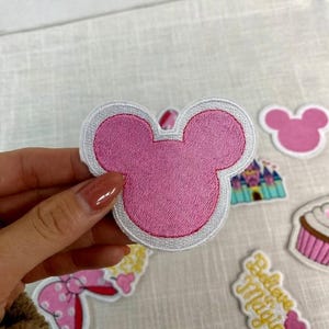 May include: Pink embroidered patch in the shape of a mouse head with a white border. Other patches include a pink mouse head, a castle, a cupcake, and a patch with the words "Believe Magic".