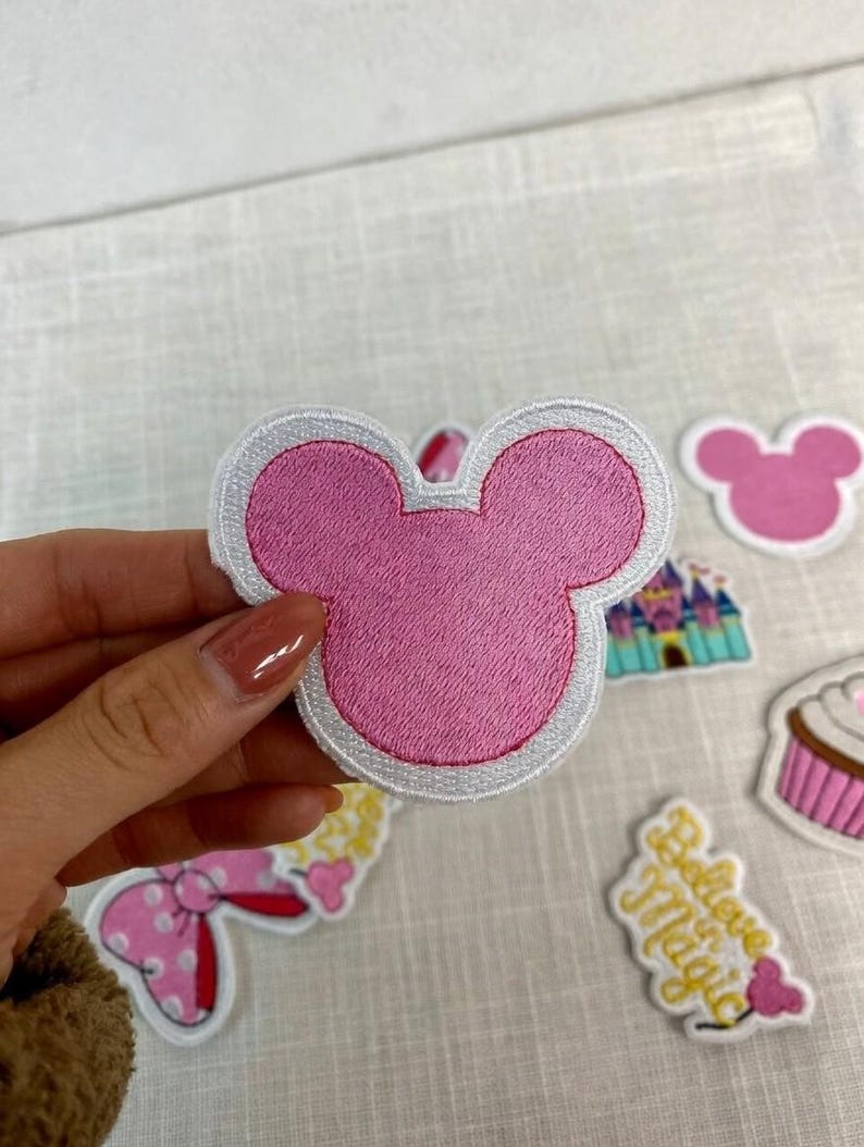 May include: Pink embroidered patch in the shape of a mouse head with a white border. Other patches include a pink mouse head, a castle, a cupcake, and a patch with the words "Believe Magic".