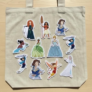 Op de afbeelding: Beige draagtas met geborduurde patches van verschillende stripfiguren. De patches tonen personages in kleurrijke jurken en outfits, waaronder een prinses met pijl en boog en een personage met een zwaard.