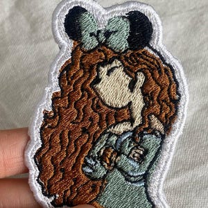 May include: Embroidered patch featuring a cartoon character with long, wavy brown hair and a green bow. The character is wearing a green dress and has a light skin tone. The patch has a white border.