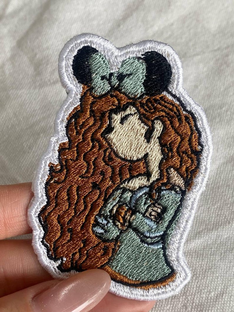 May include: Embroidered patch featuring a cartoon character with long, wavy brown hair and a green bow. The character is wearing a green dress and has a light skin tone. The patch has a white border.