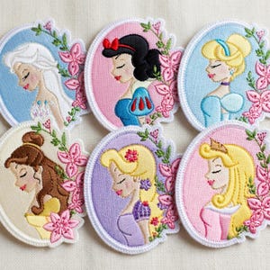 Pode incluir: Patches bordados com seis designs de princesas diferentes. Cada patch tem formato circular com um retrato de princesa, rodeado por uma grinalda floral. As princesas são retratadas em perfil, com cores suaves e detalhes delicados.