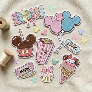 Puede incluir: Parches bordados con temática mágica. Los diseños incluyen una manzana con caramelo, palomitas de maíz, un cono de helado, una magdalena y globos con forma de Mickey Mouse. La palabra "MAGICAL" está escrita con letras coloridas. Parches adicionales tienen forma de boletos y cabezas de Mickey Mouse.