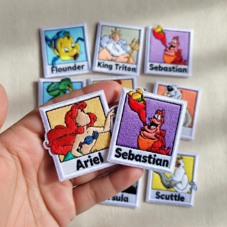May include: A collection of embroidered patches featuring characters from the animated film "The Little Mermaid." Patches include Ariel, Flounder, Sebastian, King Triton, and Scuttle. Each patch has a white border and a character name.