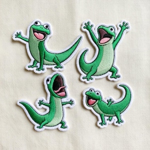 Pode incluir: Quatro patches bordados com desenhos de lagartixas de desenho animado em várias poses. As lagartixas são verdes com contornos brancos e bocas abertas, sobre um fundo creme. Cada lagartixa tem uma expressão diferente.