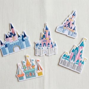 Puede incluir: Cinco parches bordados con diseños de castillos. Los parches muestran castillos con detalles en rosa, azul y dorado. Cada parche tiene un borde blanco y está sobre un fondo neutro.