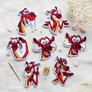 Op de afbeelding: Zeven geborduurde patches met een rode draak met een witte rand. De draak heeft een gele buik, paarse details en verschillende uitdrukkingen. De patches zijn gerangschikt op een crèmekleurig oppervlak, met een naald en draad zichtbaar.