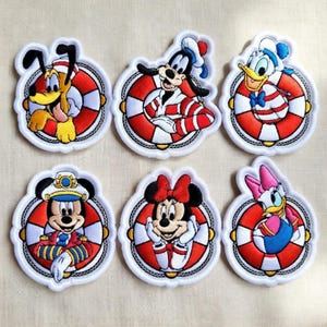 Może przedstawiać: Zestaw sześciu haftowanych naszywek z postaciami Disneya: Pluto, Goofy, Kaczor Donald, Myszka Miki, Myszka Minnie i Daisy. Każda postać umieszczona jest w czerwono-białym kole ratunkowym.