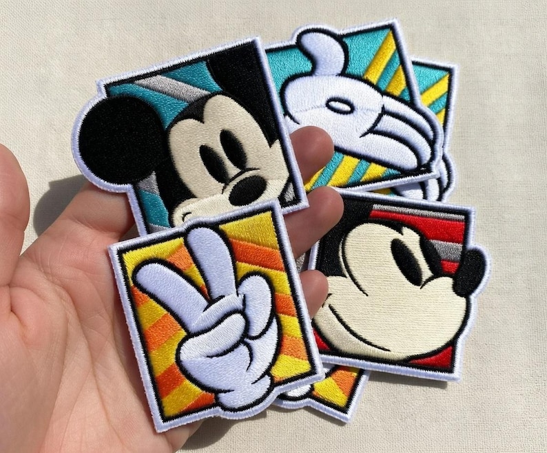 May include: A collection of embroidered patches featuring Mickey Mouse and his iconic imagery. Each patch is square with a colorful background, including a peace sign, a gloved hand, and Mickey's face. The patches are white, black, red, yellow, and blue.