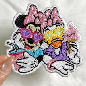 May include: Embroidered patch featuring Minnie Mouse and Daisy Duck wearing heart-shaped sunglasses. Minnie holds a drink, and Daisy holds an ice cream cone. The patch has a white border and is on a white background.