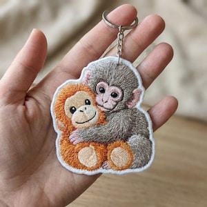 May include: Embroidered keychain featuring two cartoon monkeys hugging. One monkey is orange, the other is gray, both with smiling faces. The keychain has a silver metal ring and chain.