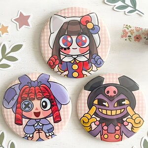 May include: Three round, colorful pinback buttons with cartoon doll designs. The buttons feature characters with large eyes, bows, and unique outfits. The background is a pink and white gingham pattern, with decorative stickers and floral tape.
