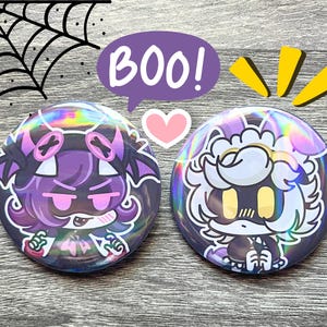 May include: Two round, holographic buttons with cartoon characters. One button features a purple character with pink accents, the other a white and yellow character. A purple speech bubble says "BOO!" with a pink heart and yellow accents.