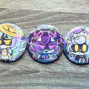 May include: Three round, holographic buttons with cartoon character designs. The buttons feature characters in white, purple, and gold with angel wings, halos, and other decorative elements. The buttons are on a gray wooden surface.
