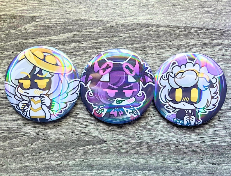 May include: Three round, holographic buttons with cartoon character designs. The buttons feature characters in white, purple, and gold with angel wings, halos, and other decorative elements. The buttons are on a gray wooden surface.