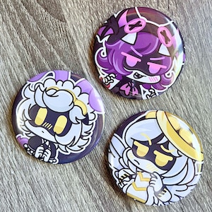 May include: Three round buttons with cartoon character designs. The buttons feature characters in purple, white, and gold with black accents. The buttons are on a wooden surface.
