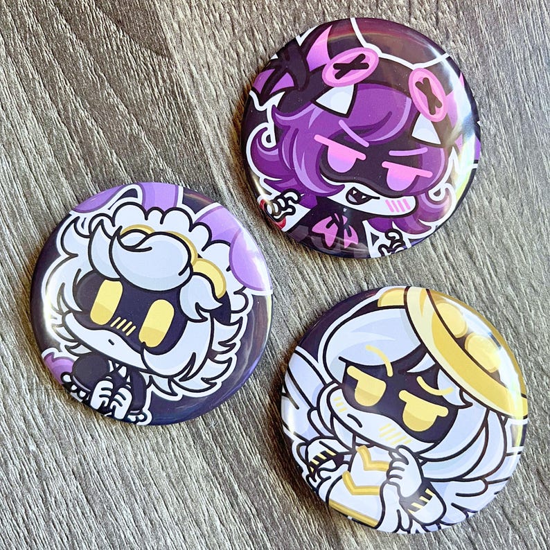 May include: Three round buttons with cartoon character designs. The buttons feature characters in purple, white, and gold with black accents. The buttons are on a wooden surface.
