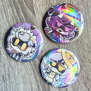 May include: Three round, holographic buttons with cartoon character designs. The buttons feature characters in purple, pink, and gold with black outlines. The buttons have a shiny, iridescent finish, reflecting a rainbow of colors.