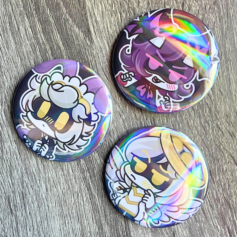 May include: Three round, holographic buttons with cartoon character designs. The buttons feature characters in purple, pink, and gold with black outlines. The buttons have a shiny, iridescent finish, reflecting a rainbow of colors.