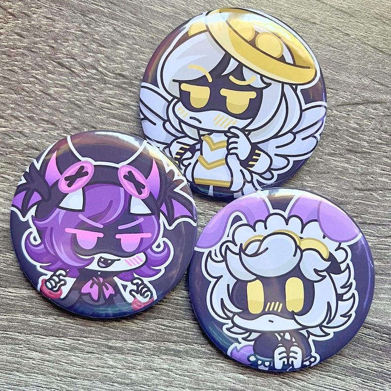 May include: Three round, colorful pinback buttons featuring anime-style character designs. The buttons showcase characters with angel wings, halos, and other unique features, in shades of purple, white, and yellow. The buttons are on a wooden surface.