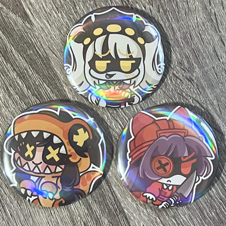 May include: Three holographic round buttons with cartoon character designs. The buttons feature characters with various expressions and outfits, including a dinosaur, a character with a knife, and a character with a crown.