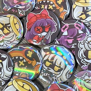 May include: A collection of round, holographic-effect buttons with cartoon character designs. The buttons feature various characters with different expressions and colors, including red, purple, and orange. Some buttons have a knife or cross design.