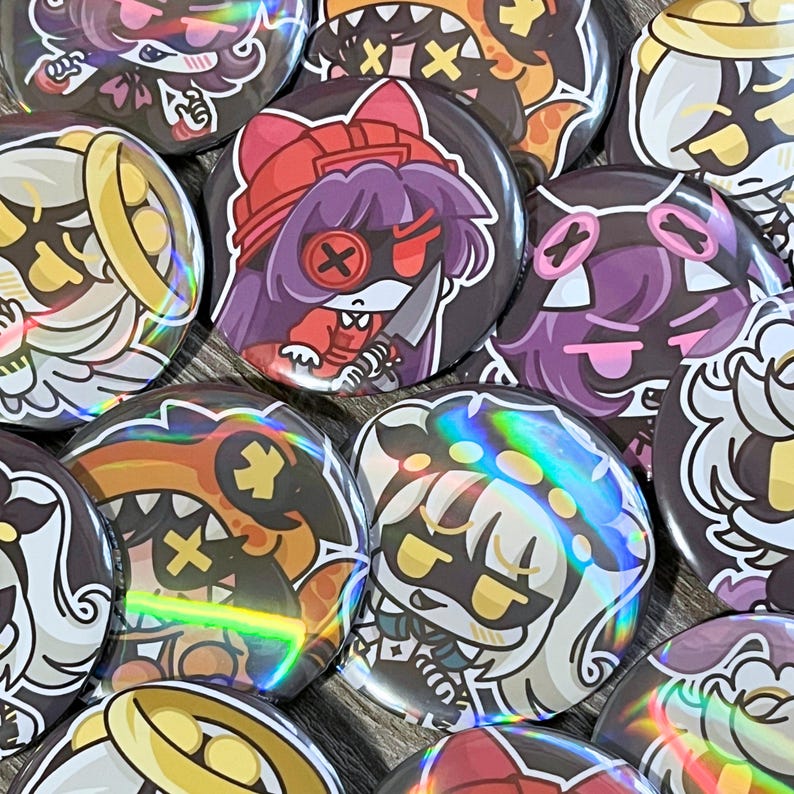 May include: A collection of round, holographic-effect buttons with cartoon character designs. The buttons feature various characters with different expressions and colors, including red, purple, and orange. Some buttons have a knife or cross design.