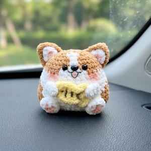 May include: A small, round, crocheted corgi plush toy featuring tan and white yarn, black plastic eyes, a small embroidered nose and mouth, pink blush accents on the cheeks, and a yellow crocheted fish-shaped snack held in its front paws.