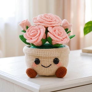 May include: A crocheted flower pot with a smiling face, holding pink roses with green leaves. The pot is beige with black button eyes and brown feet. The flowers are a soft pink color.