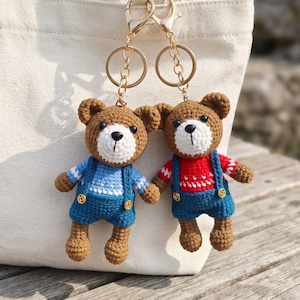 Peut inclure: Deux porte-clés ours en crochet marron avec des fermoirs en métal doré. Un ours porte un pull bleu clair et une salopette bleu foncé, tandis que l'autre porte un pull rouge à motifs blancs et une salopette bleu foncé. Tous deux ont un museau blanc.